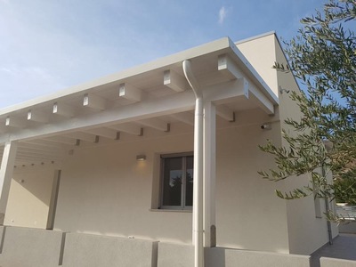 Vista frontale di una tettoia in legno lamellare Abete impregnata bianco con copertura finale in alluminio aggraffato complete di gronde e accessori, realizzata a Trapani (TP).